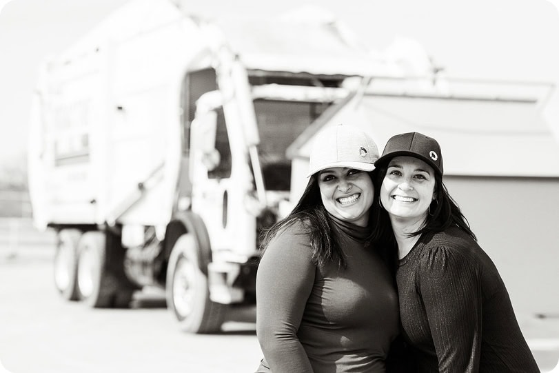 Two women in safety helmets smiling in front of waste management truck.