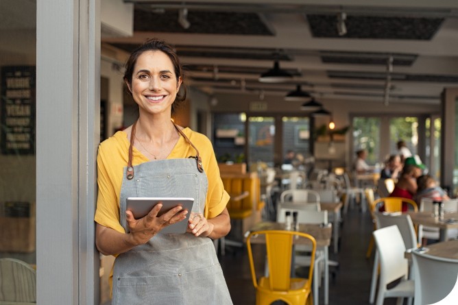 cafe owner in front of busy cafe holding ipad and smiling at camera