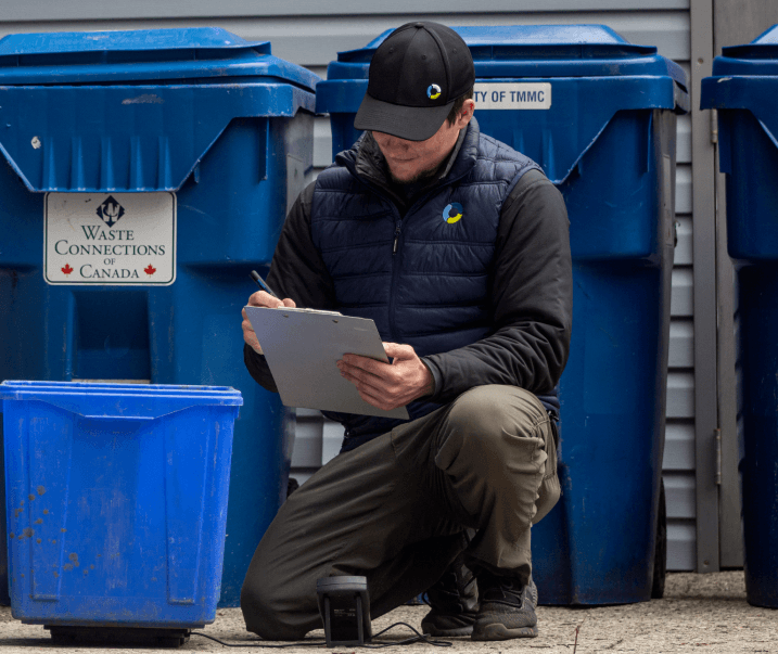 waste auditor smiling in front of recycling bins while weighing bin and holding checklist