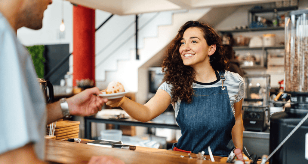 Woman handing over a coffee to a customer at a cafe counter, promoting waste reduction.